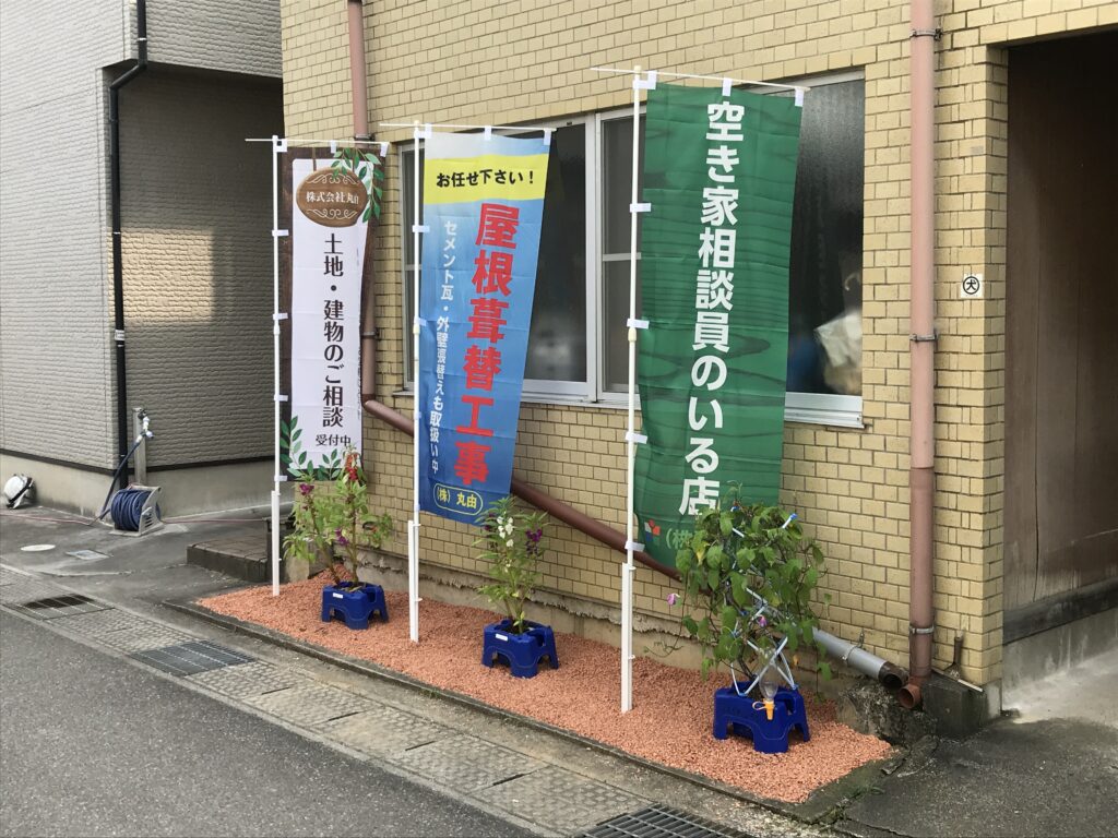 株式会社丸由の会社外観写真。屋根葺替工事や空き家相談を案内するのぼり旗が設置された入口。