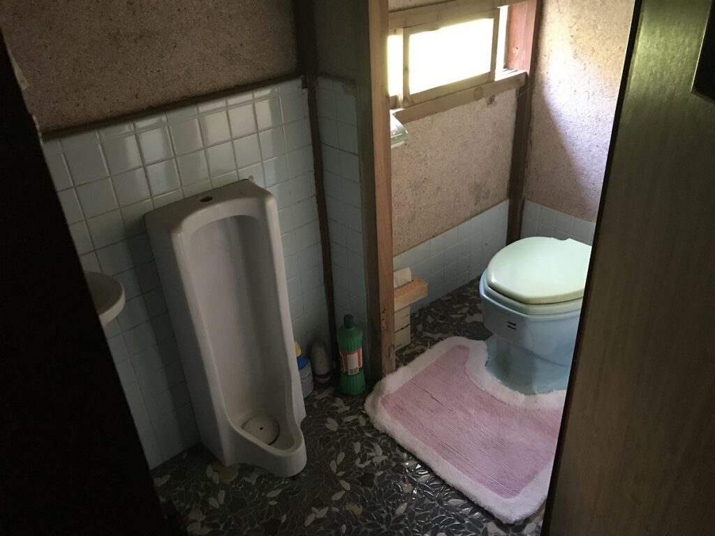 滋賀県犬上郡の空き家「円城寺邸」のトイレ。洋式便器と小便器が並ぶ昭和期の設備。タイル壁とモルタル床、採光窓あり